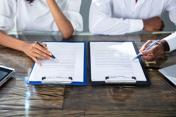Two people in white shirts sit at a wooden desk, reviewing and pointing at documents on clipboards with pens.