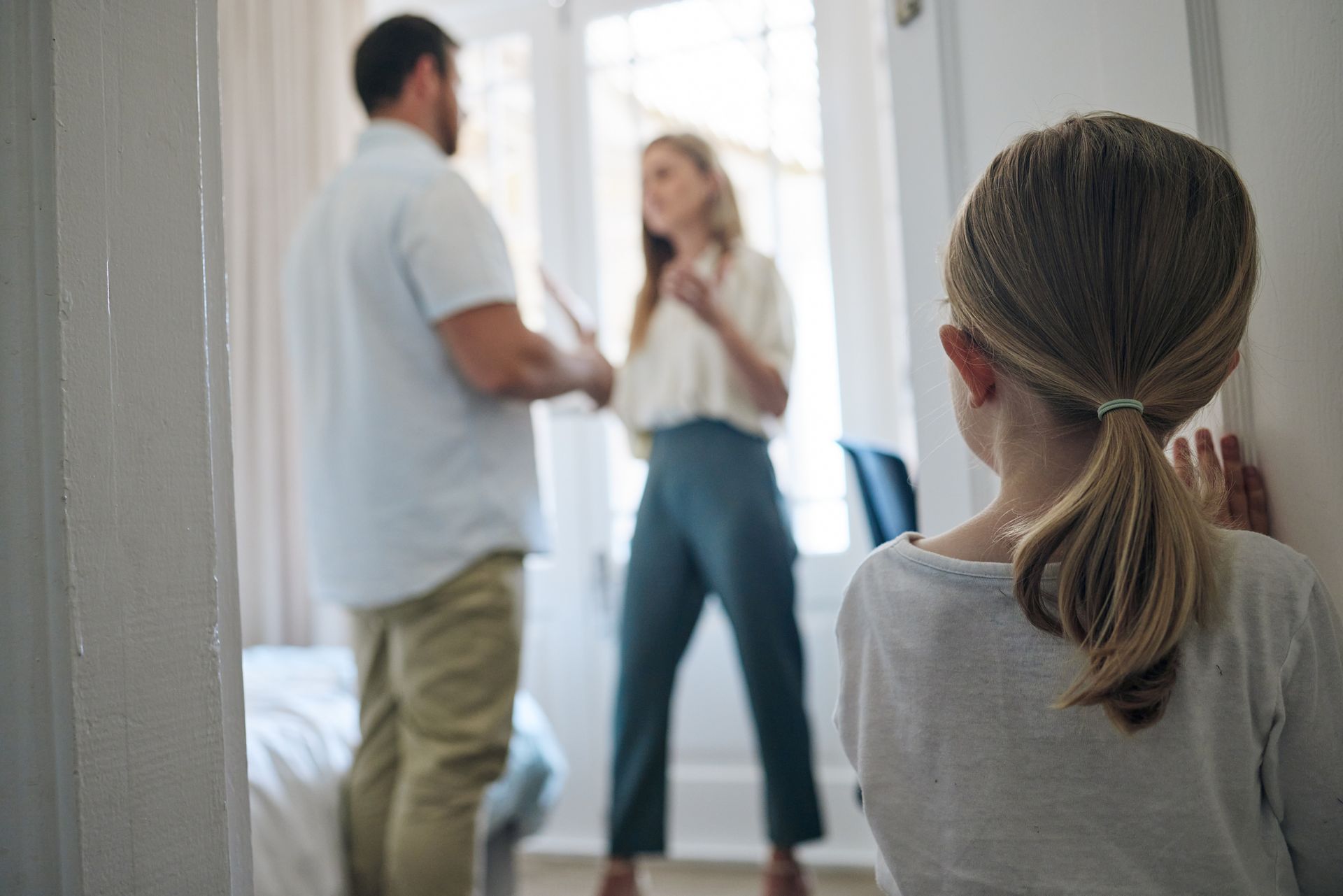 A young girl watches two adults arguing in a bright room, seen from behind.