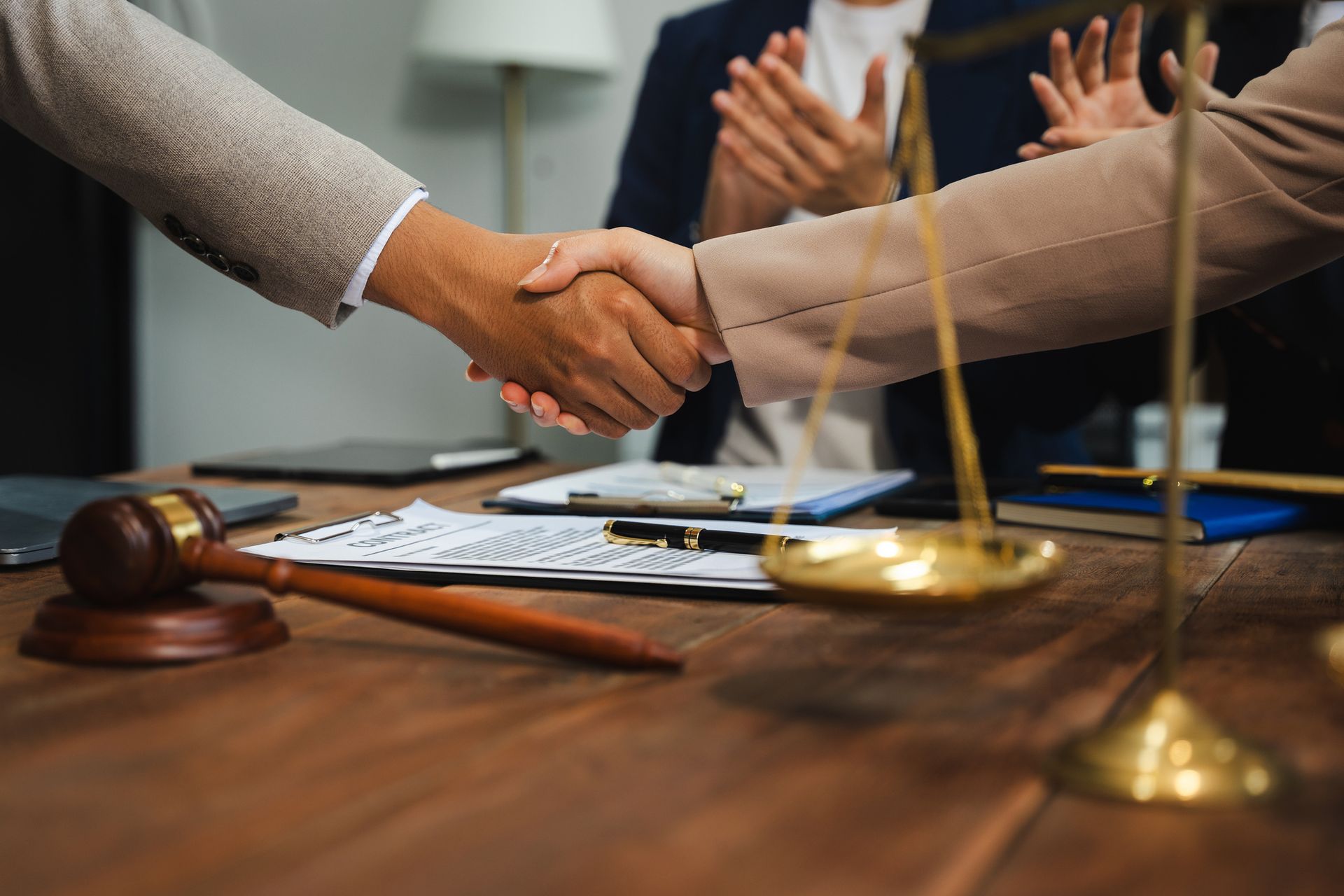 Two people shaking hands over a wooden desk with a wooden gavel and a gold scale of justice in the foreground.