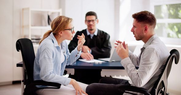 Two people argue while seated across from each other in an office, with a third person observing in the background.