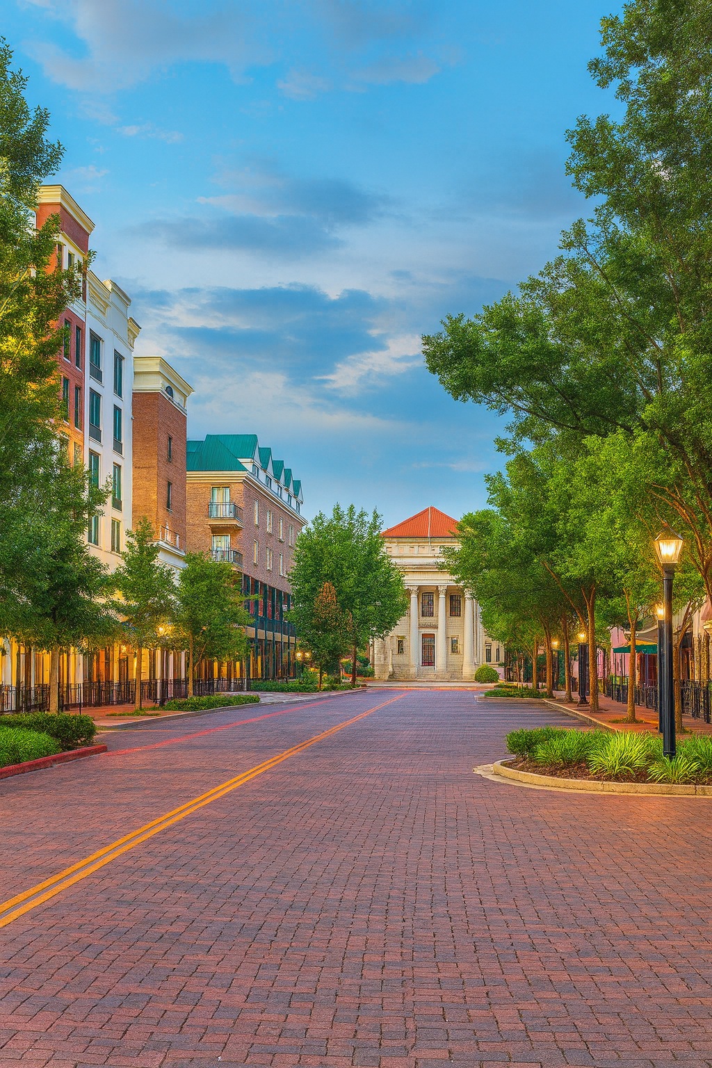 Brick street lined with buildings and trees, leading to a neoclassical building under a blue sky.