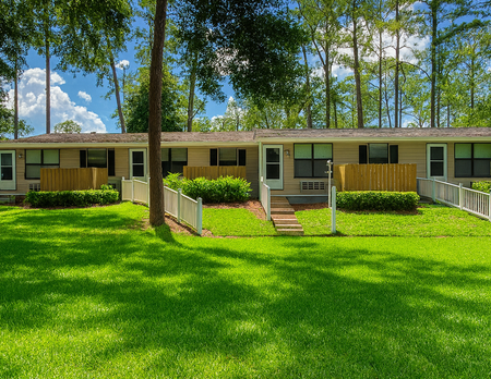 Row of beige apartments with green lawns, white picket fences, and trees.