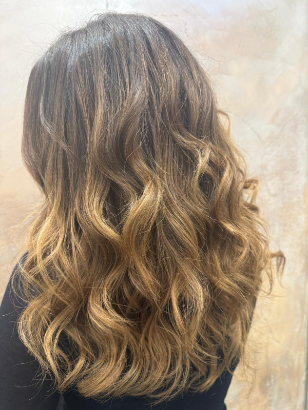 Vista posteriore di lunghi capelli castani ondulati con riflessi biondi balayage, portati sciolti sopra un top nero.