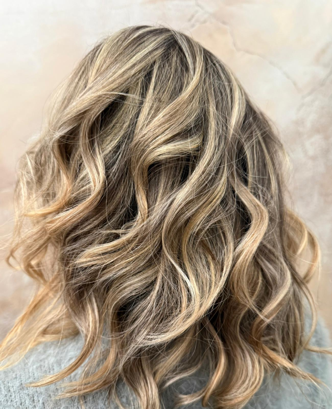 Vista posteriore di capelli ondulati lunghi fino alle spalle, con riflessi biondi multitonali e radici scure.