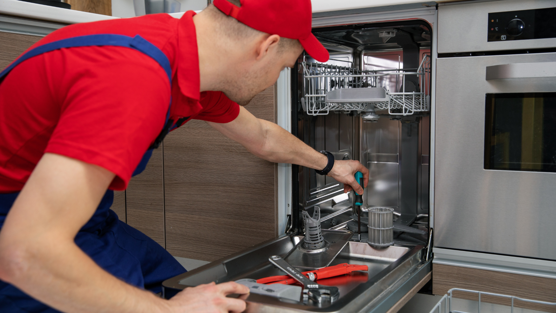 Man in red shirt and cap repairing a dishwasher, using tools, indoors.