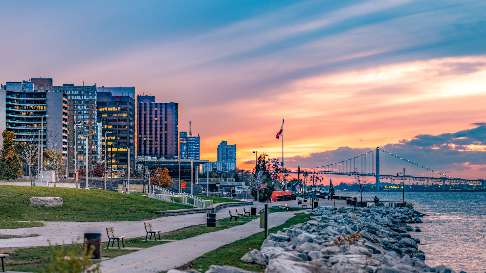 City skyline at sunset, buildings next to a waterfront with a walkway, orange and blue sky.