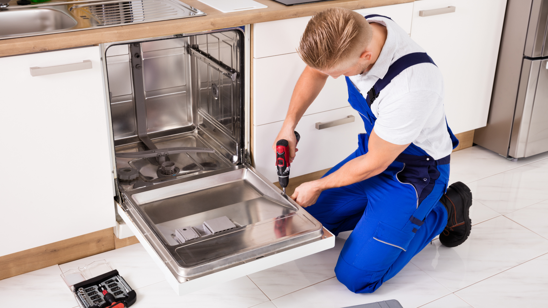 A person in blue overalls fixing a dishwasher with a power drill in a kitchen setting.