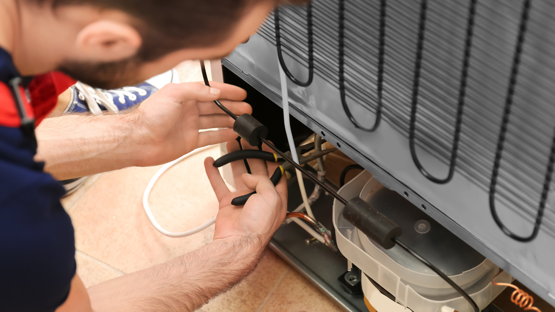 Person repairing refrigerator, using pliers to manipulate wires near the compressor and cooling coils.