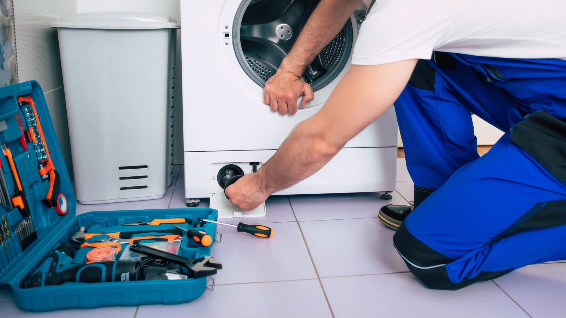Person in blue overalls repairs washing machine in a bathroom. Toolbox with tools sits open nearby.