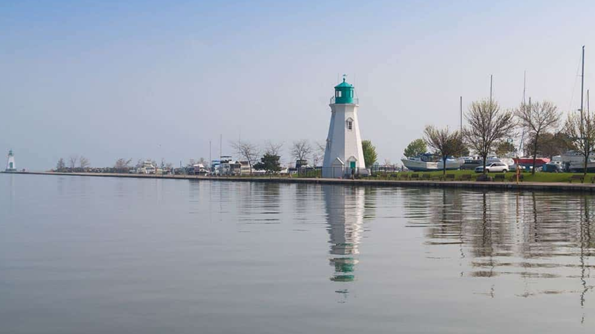 Lighthouse on calm water, reflected. Green and white tower. Distant boats and shoreline under a blue sky.