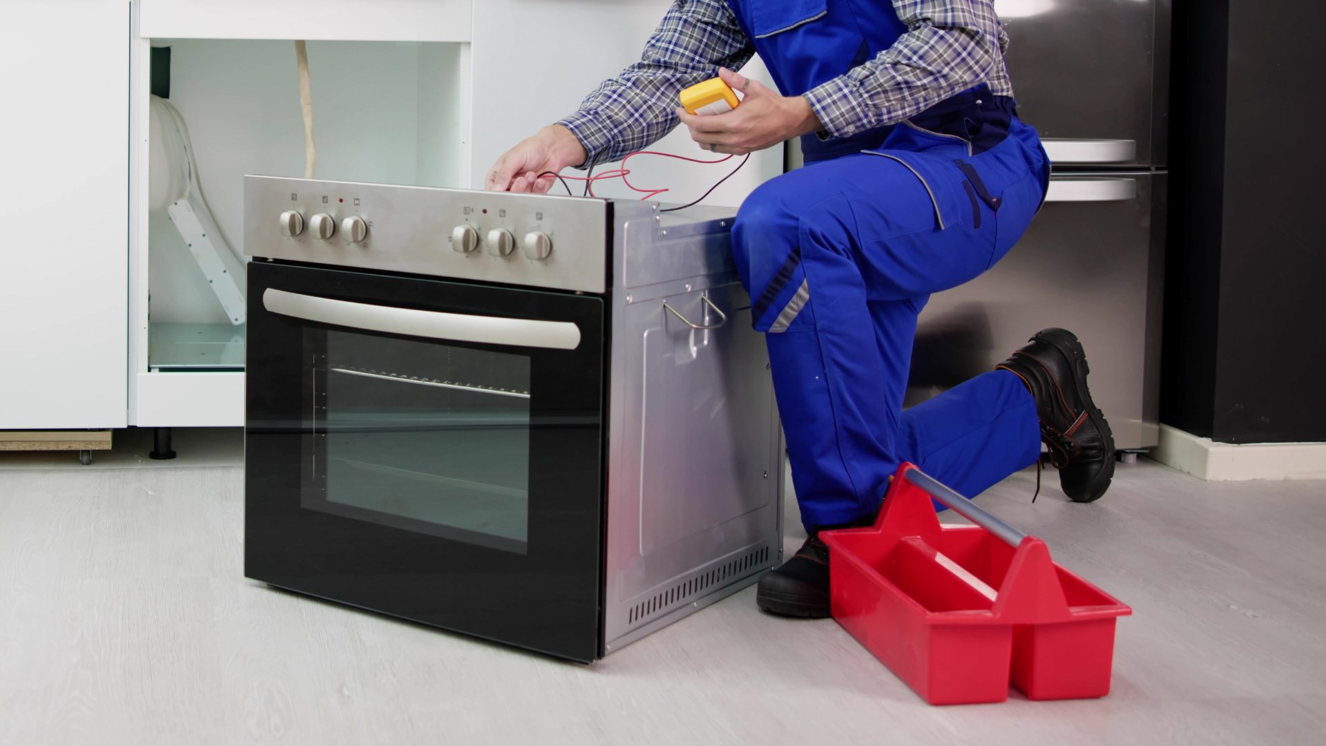 Technician in blue overalls repairing an oven, kneeling beside a red toolbox.