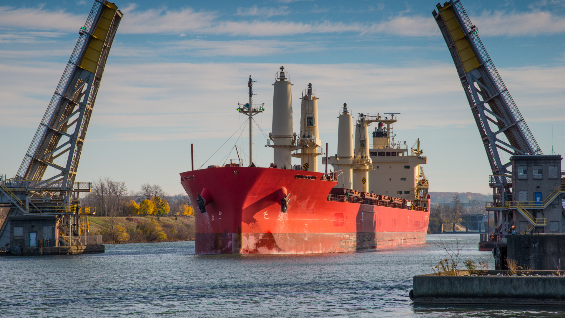 Red cargo ship sailing through open drawbridge, on a sunny day.