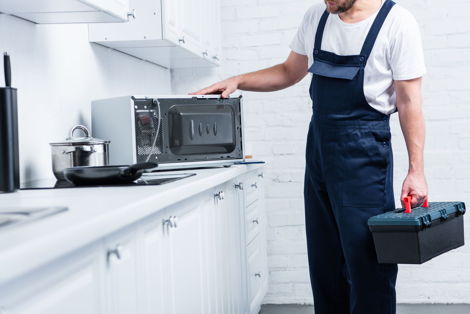 A repairman in blue overalls, holding a toolbox, examines a microwave in a white kitchen.