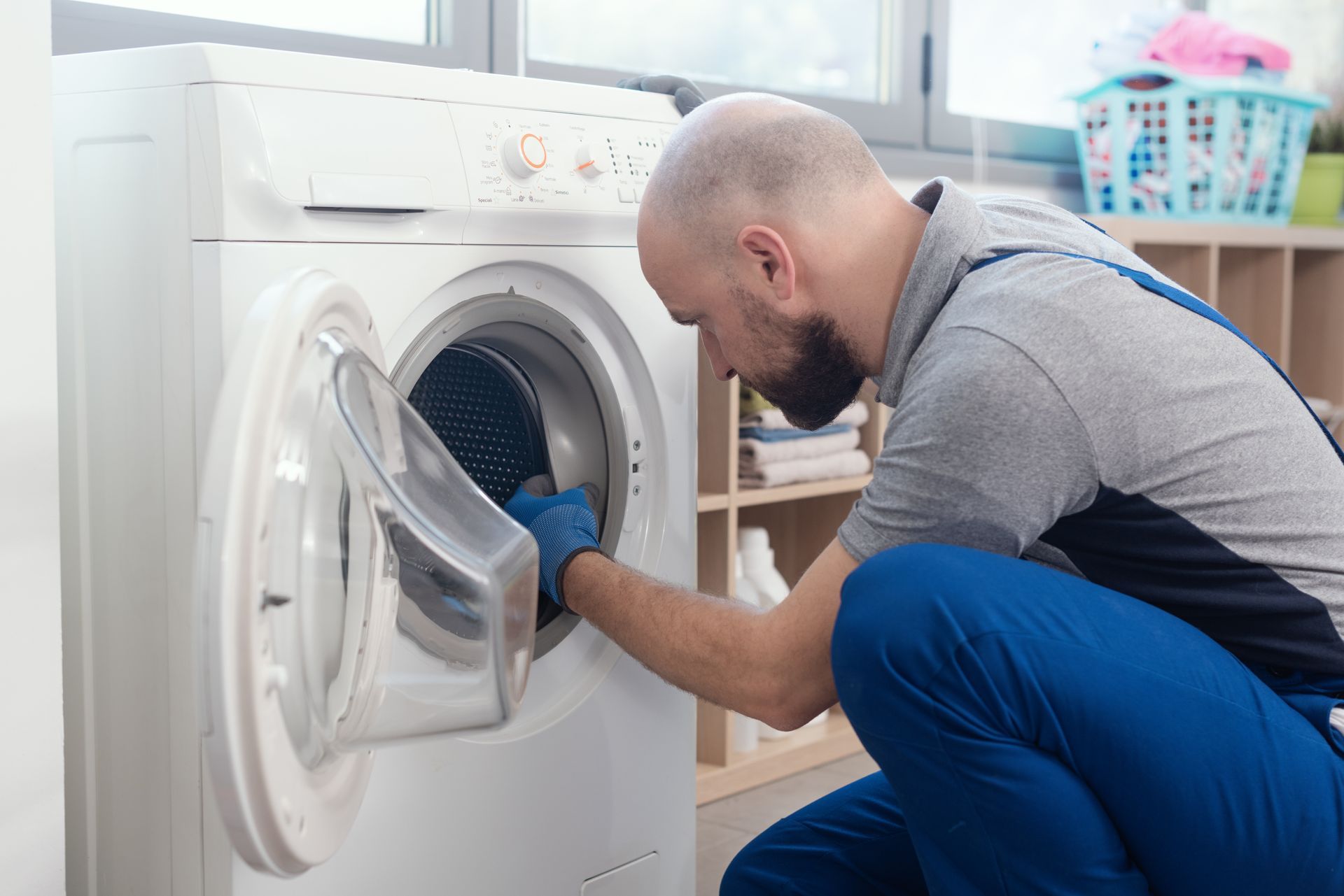 Man in blue overalls repairs washing machine, opening the door in a laundry room.