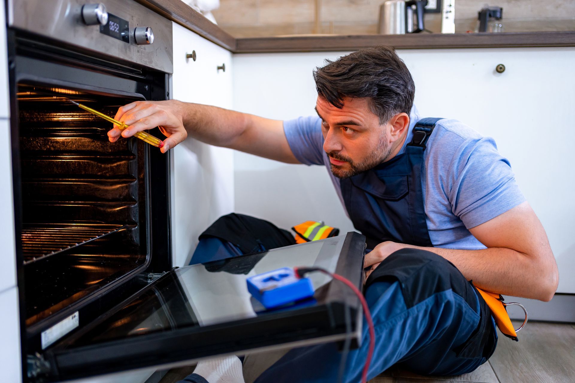 Man inspecting an oven with a tool. He kneels near the appliance in a kitchen, using a multimeter.