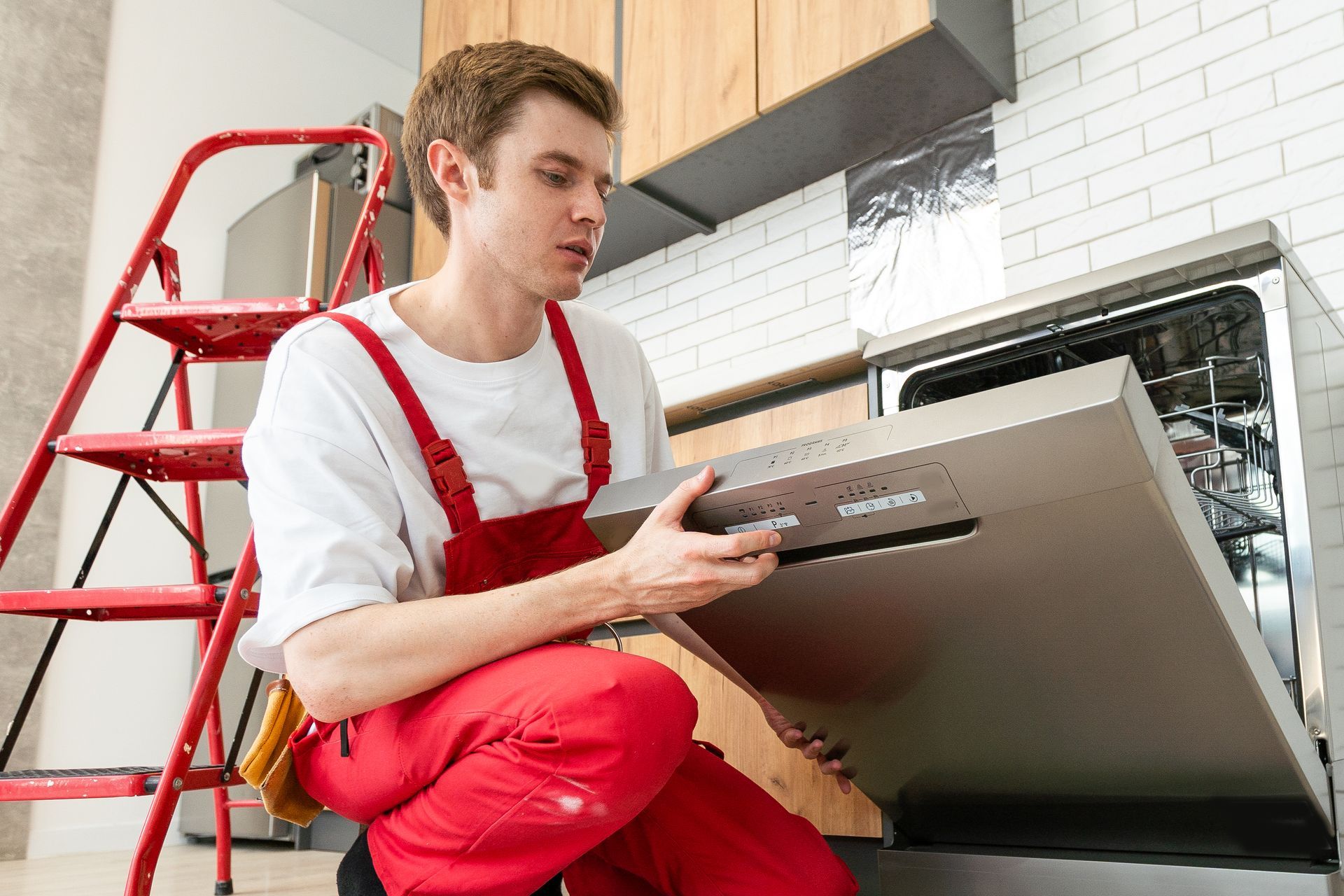 Man in red overalls installing a dishwasher in a kitchen; ladder nearby.