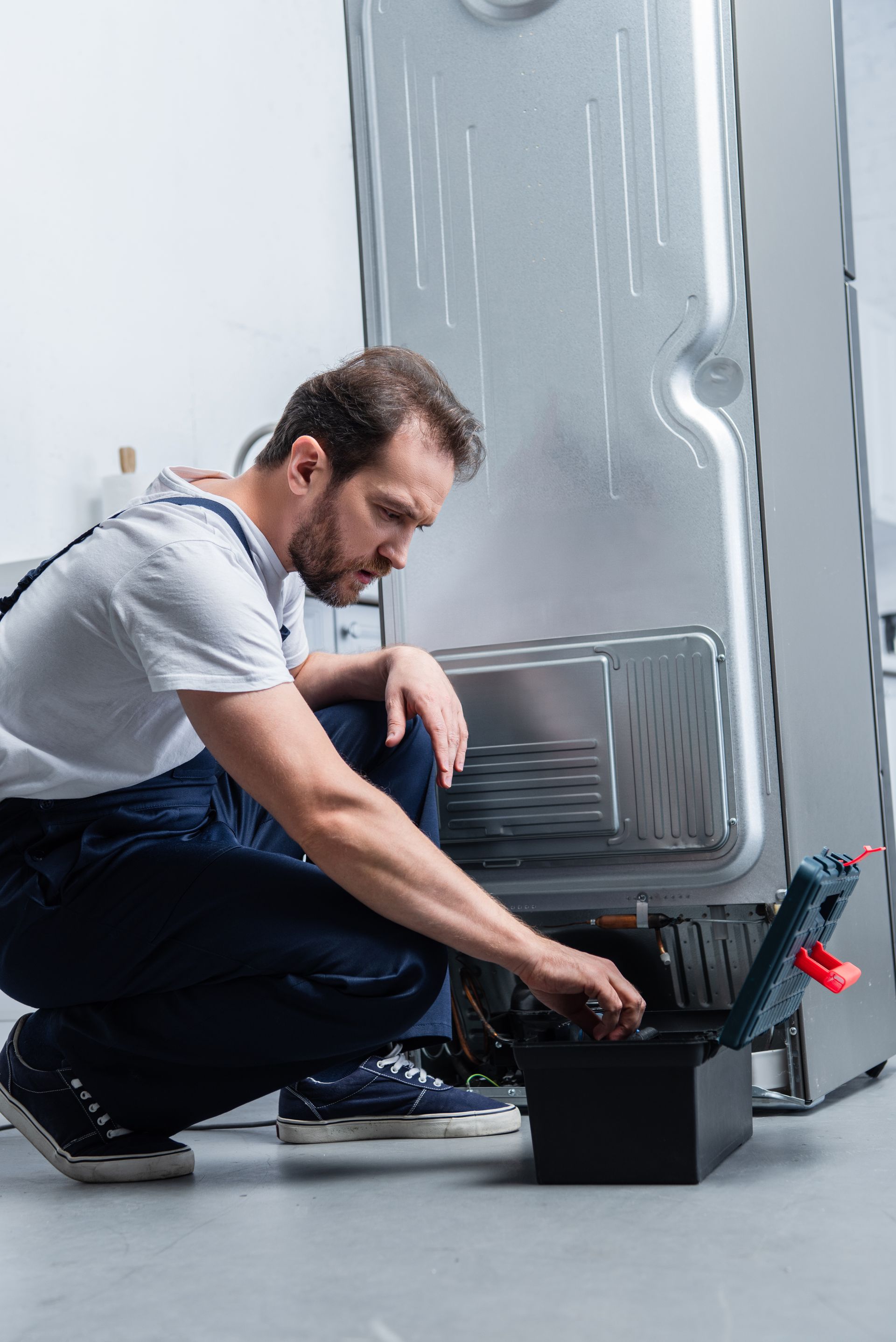 Man in blue overalls kneels, inspecting appliance with toolbox. Silver fridge in a light room.