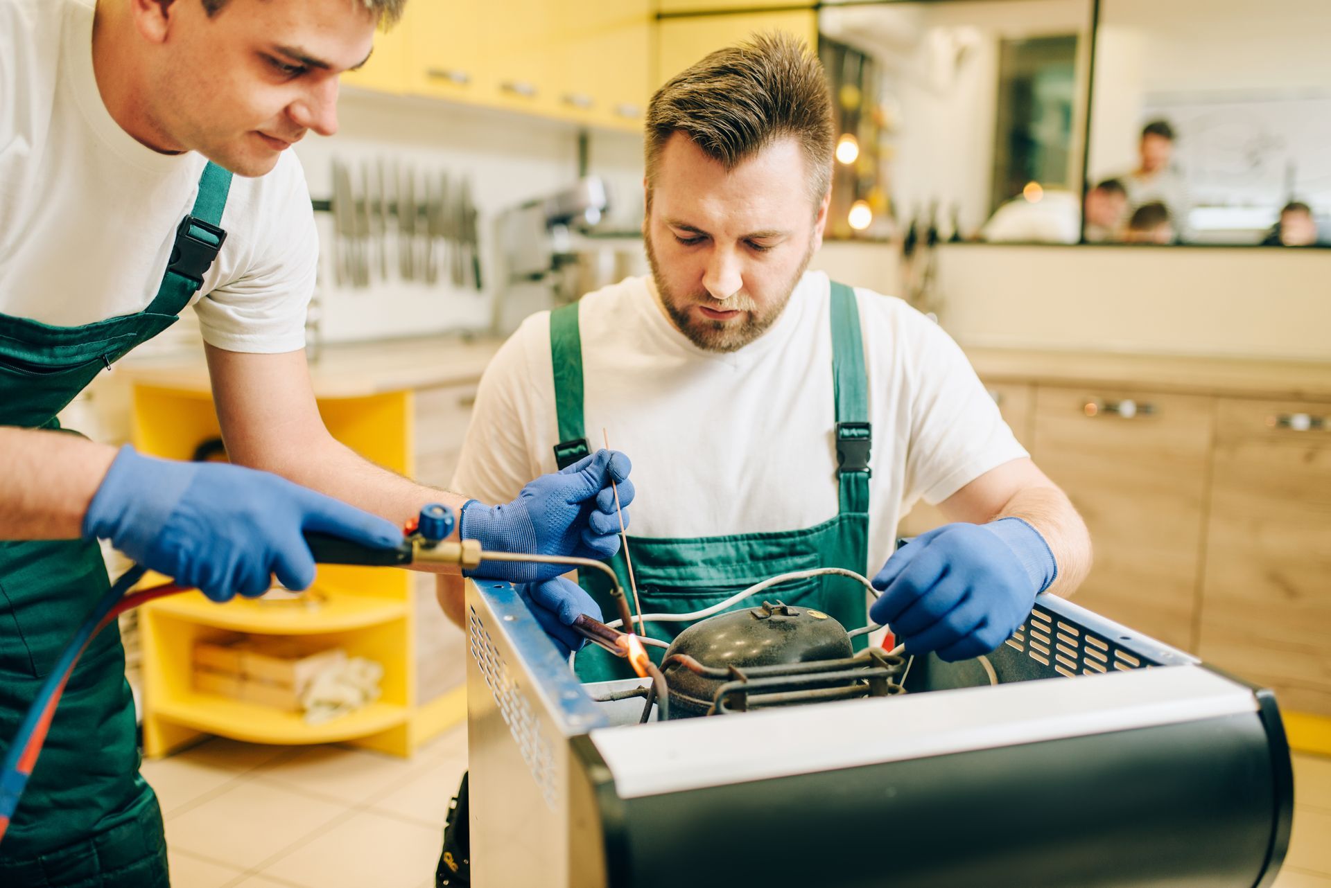 Two technicians in blue gloves work on a refrigerator using a blowtorch in a kitchen.