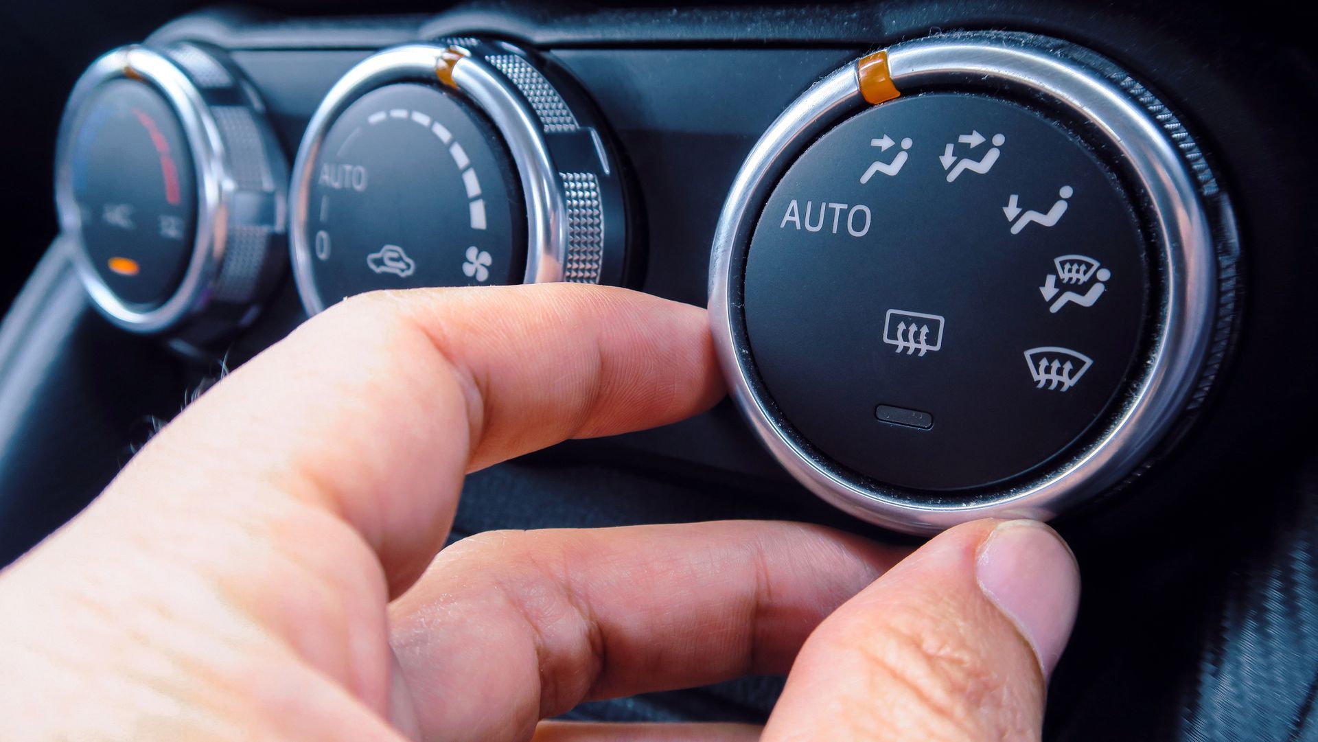 Primer plano de una mano ajustando los diales del climatizador del salpicadero negro de un coche con iconos blancos.