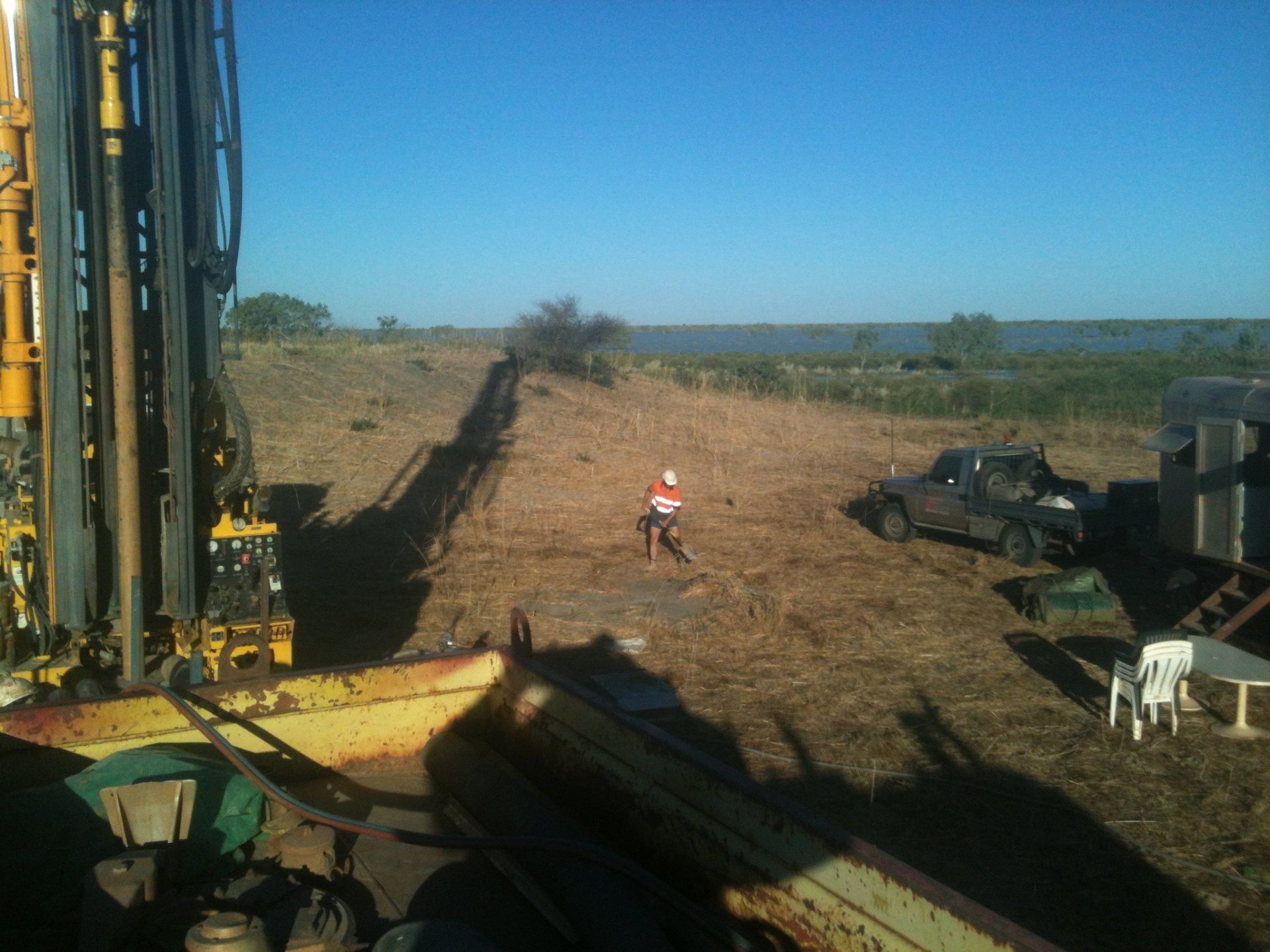 Drilling rig in field, person in orange shirt, small trucks, blue sky, water in the distance.