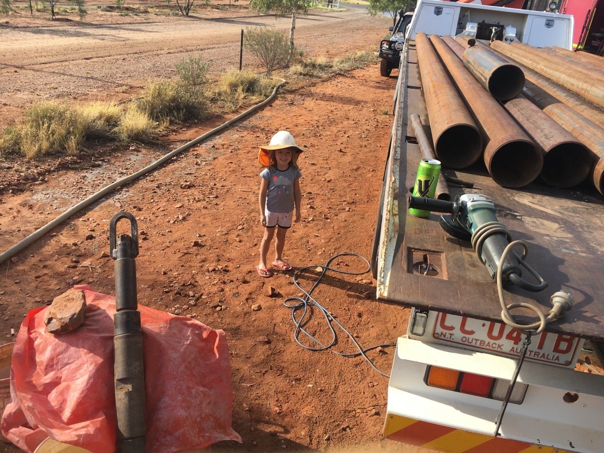 A child wearing a hard hat stands near a truck with pipes and tools on a red dirt road.