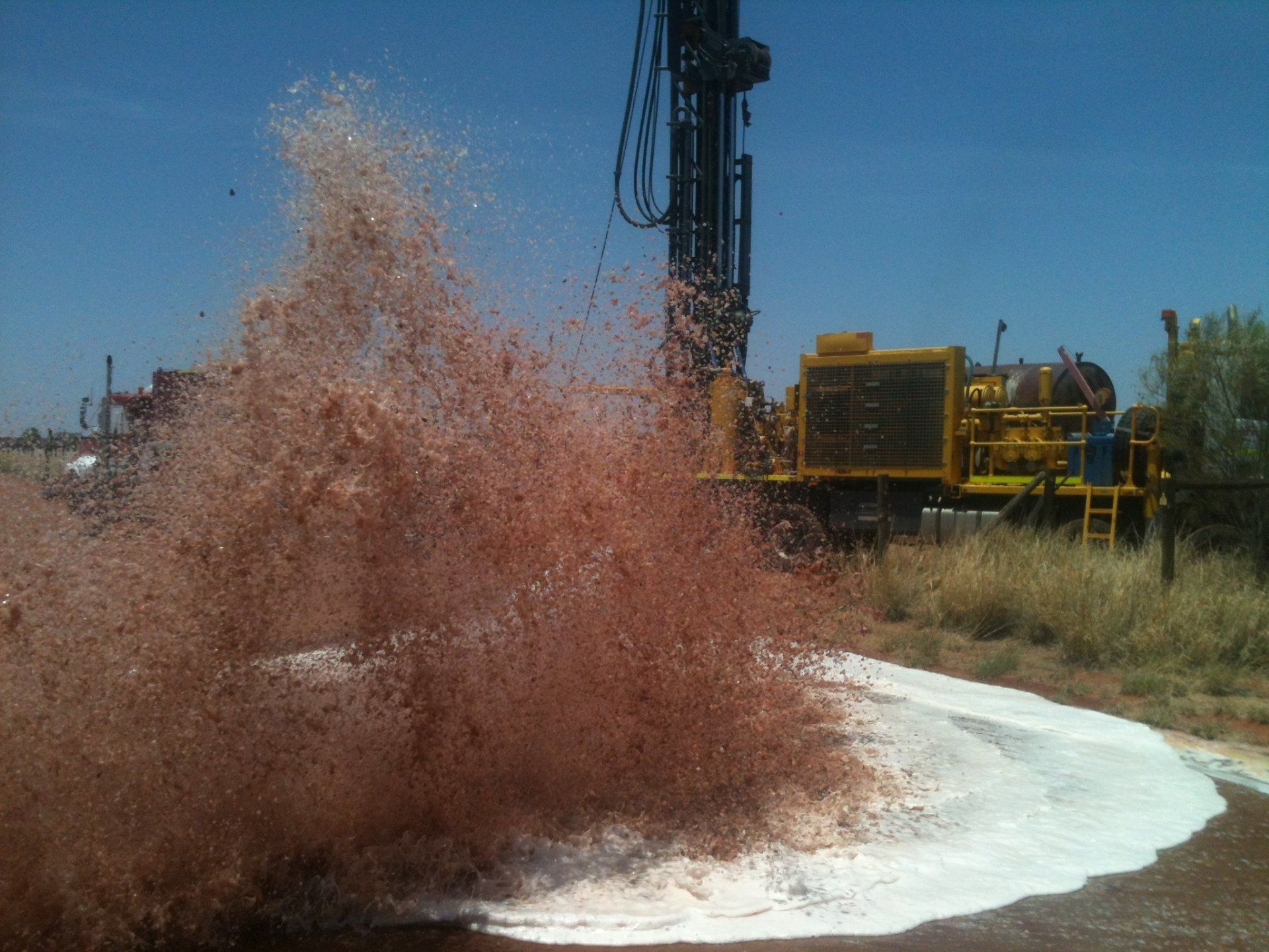 A well drilling rig spews red-brown mud onto the ground, with a blue sky in the background.