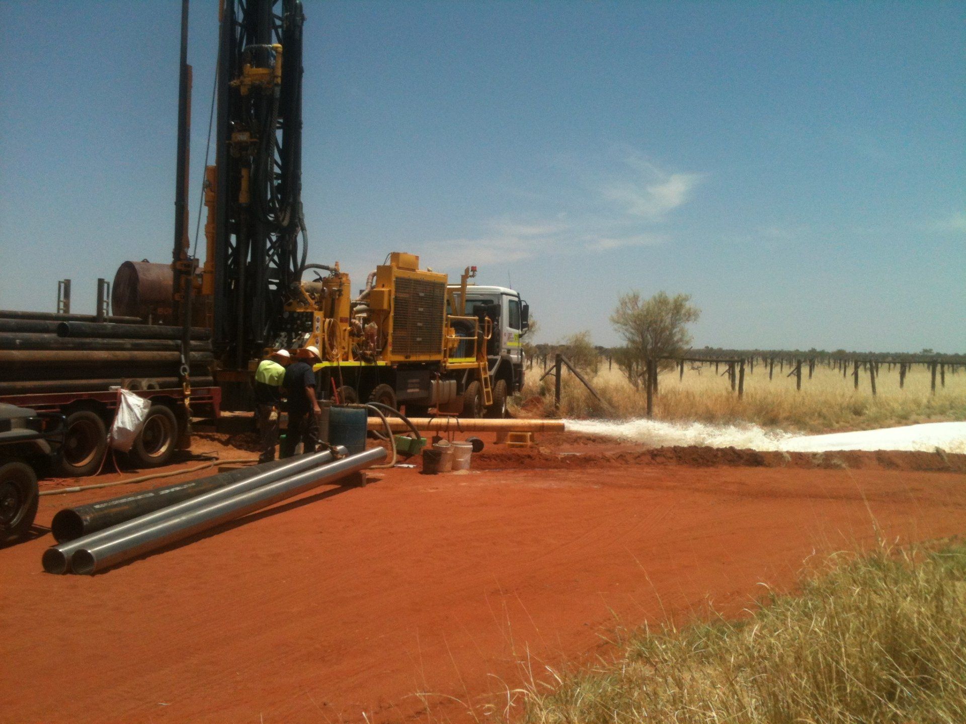 Drilling rig on red earth, workers assembling pipes. Sunny day, field in background.