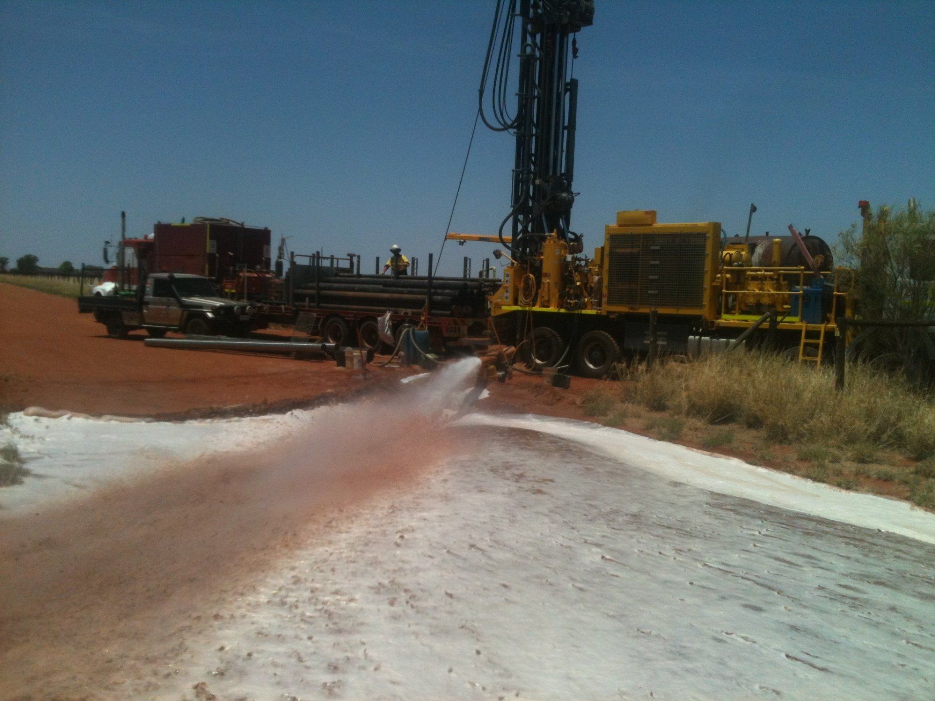 Drilling rig sprays water on a dusty road. A truck is parked nearby on a sunny day.