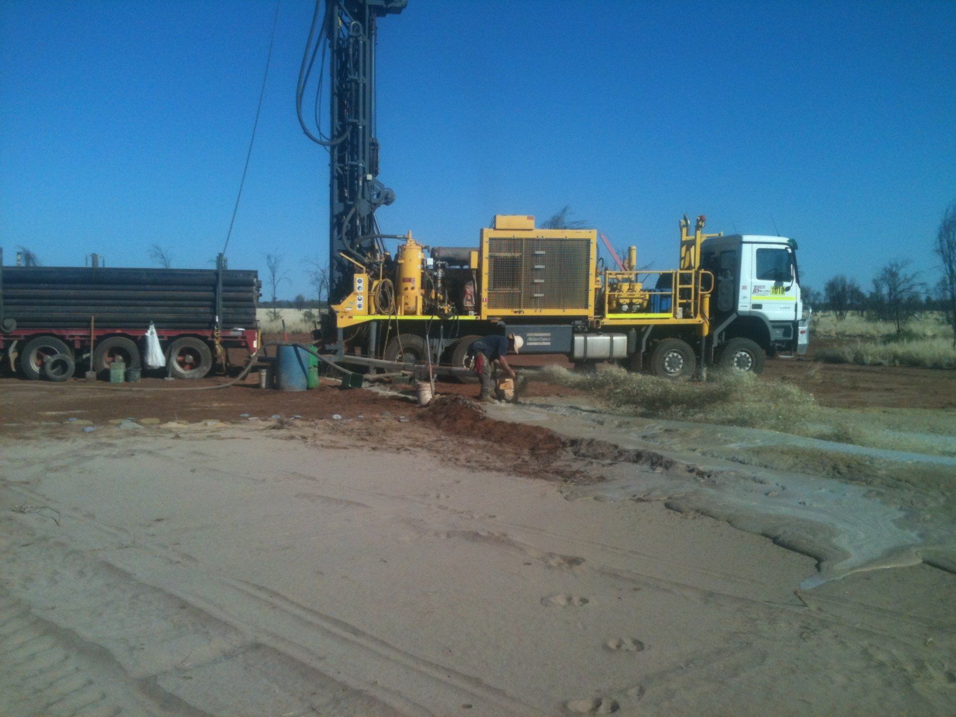 Yellow drilling rig on truck, drilling in sandy area with black pipes on a trailer.