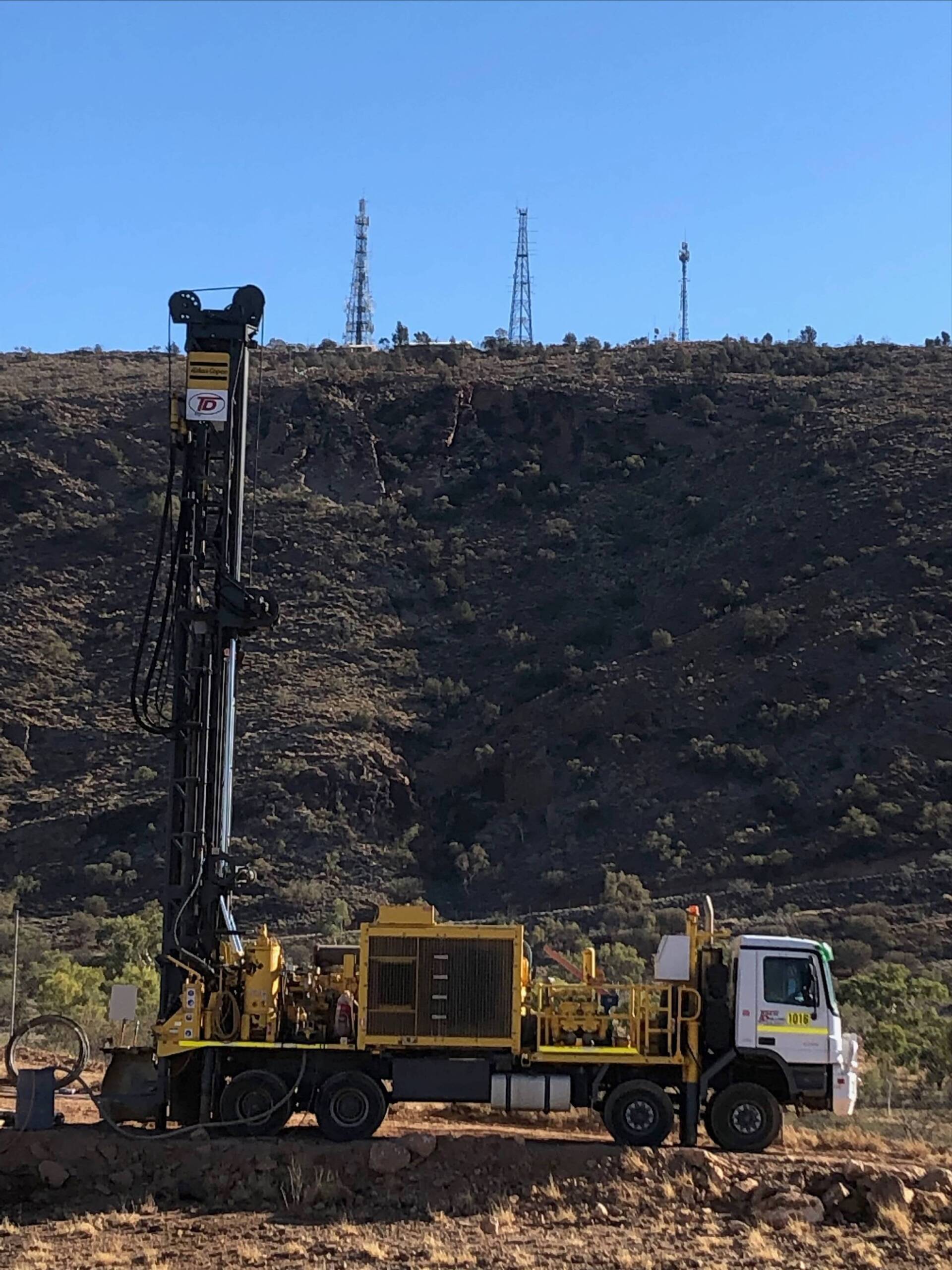 Drilling rig on wheels, on a brown and rocky hillside, under a clear blue sky.
