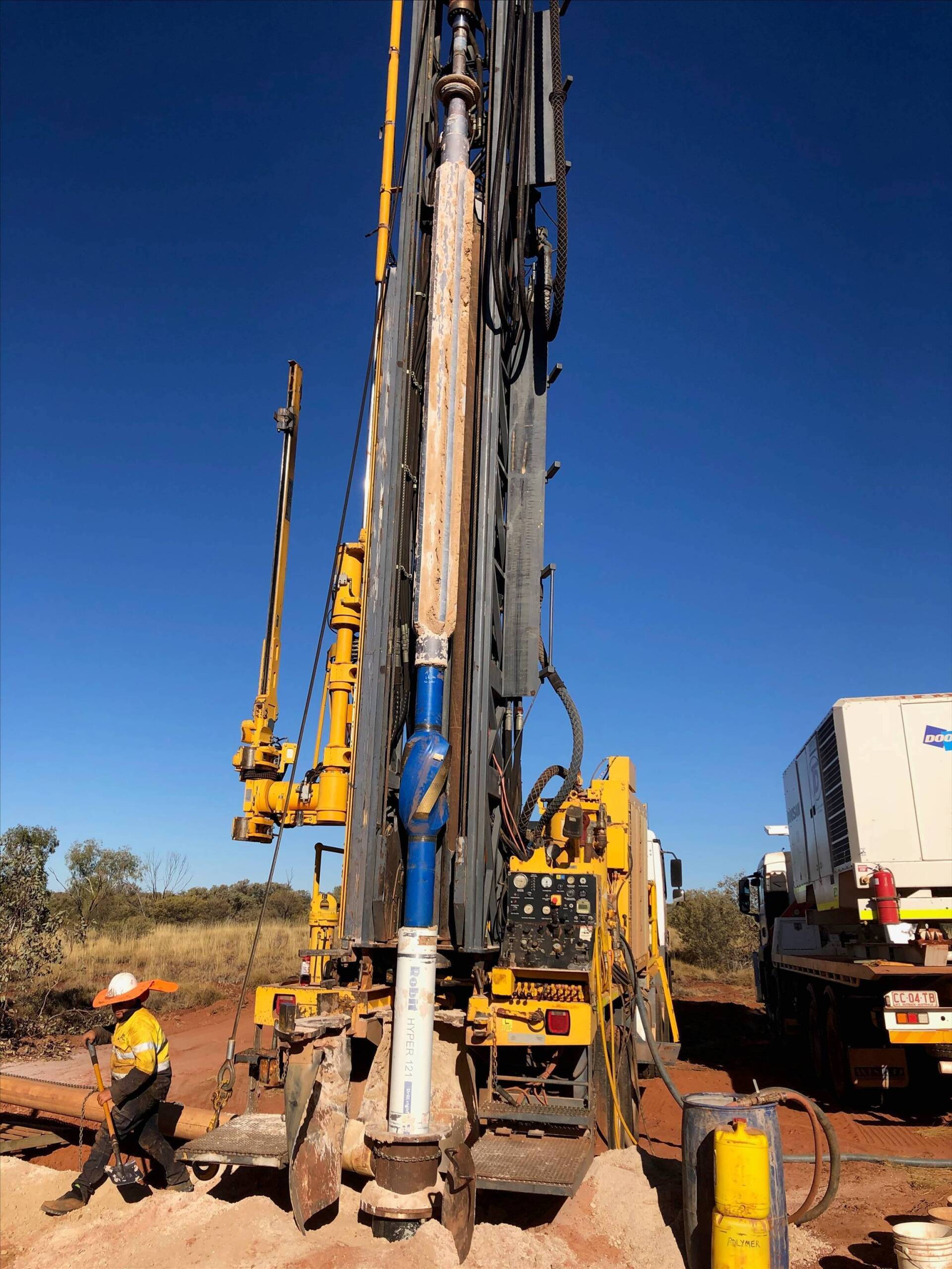 Yellow drill rig in an outdoor setting, drilling. A worker in safety gear is on the left.