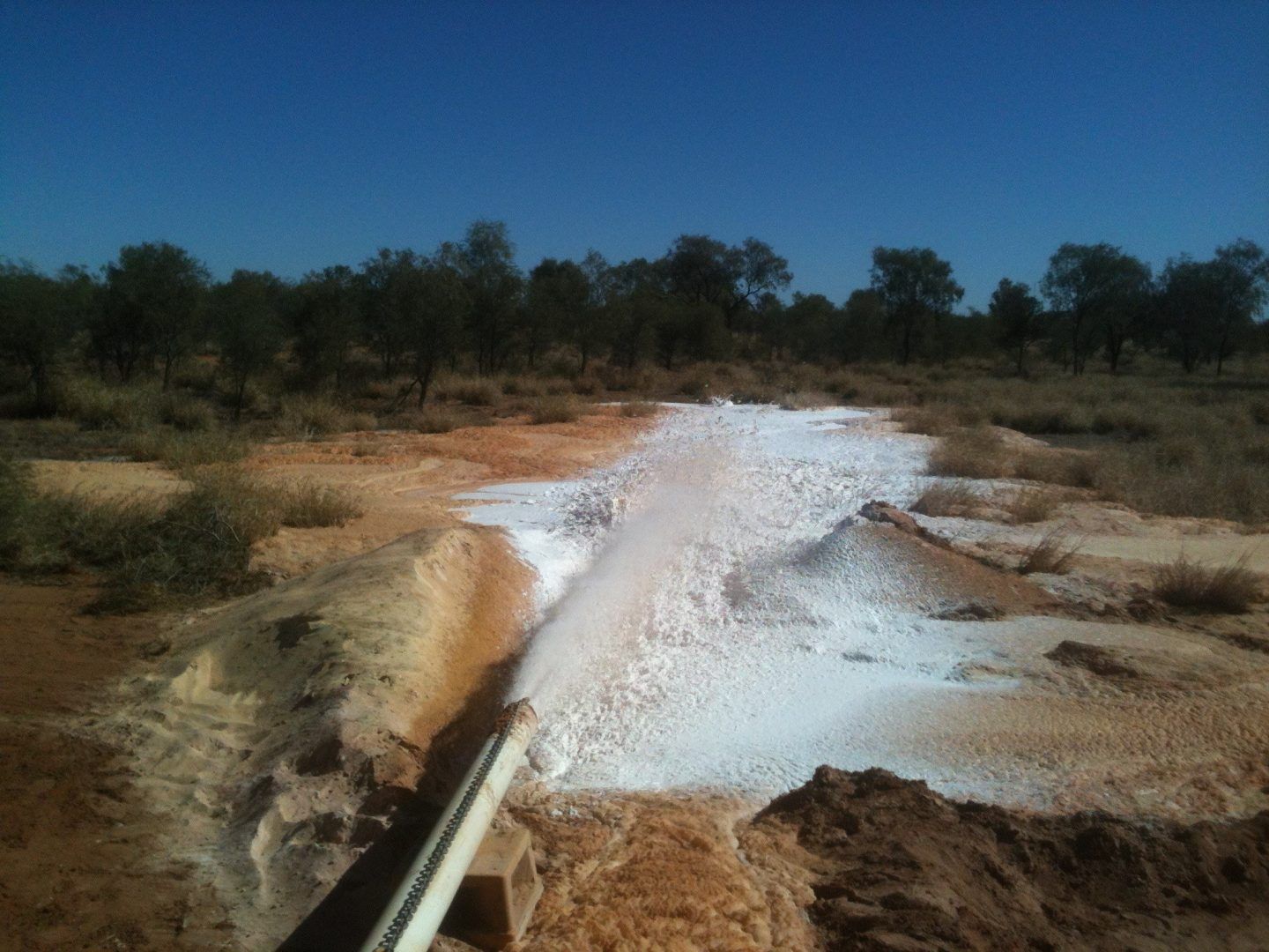 Water gushing from a pipe, creating a foamy, white pool on reddish-brown earth, in a sunny, outdoor setting.