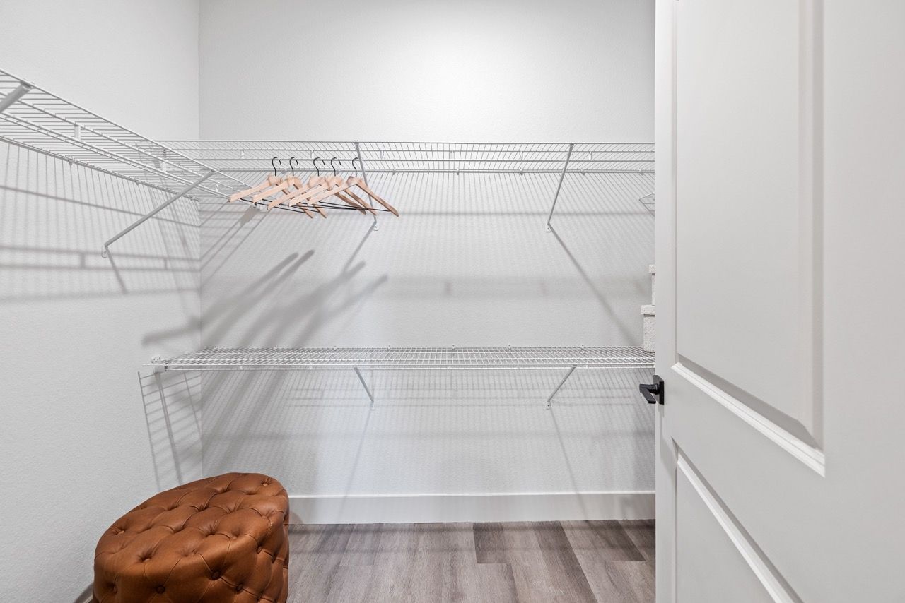 Empty walk-in closet with white wire shelves, wooden hangers, and a brown tufted ottoman.