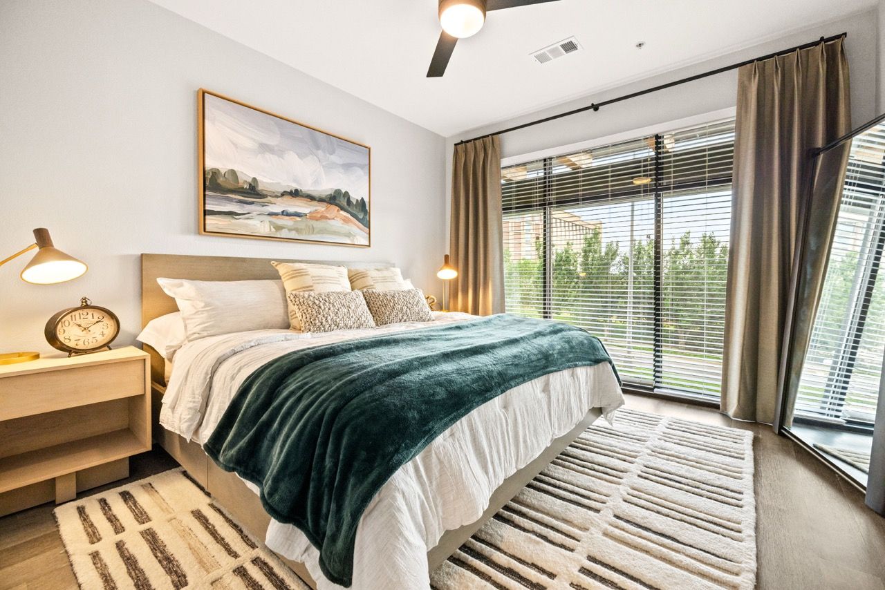Modern apartment bedroom with a large window, blinds, and a teal throw on the bed.