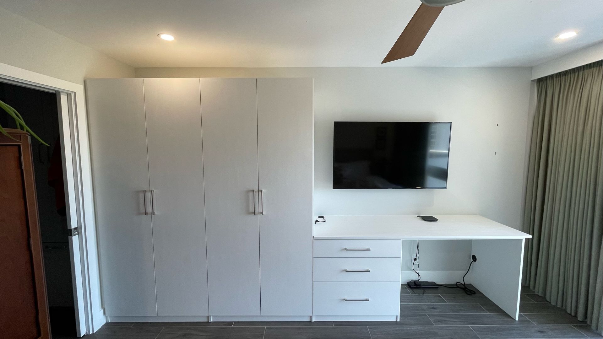 A bedroom with white cabinets , a desk , a flat screen tv and a ceiling fan.