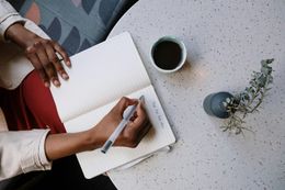 Person writing in notebook with coffee and plant on a table.