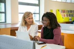 Woman helping another woman with laptop, seated in library.