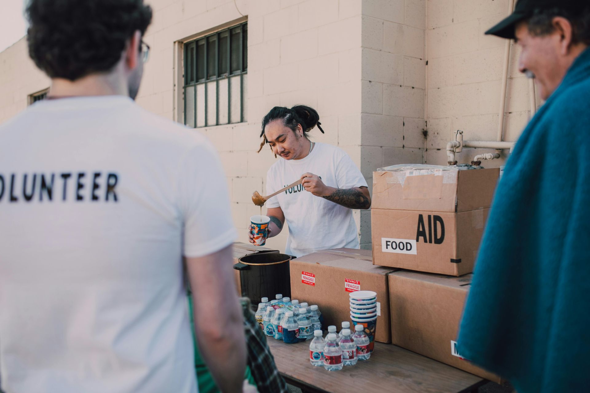 Volunteers serving food outdoors from a pot, alongside boxes labeled 