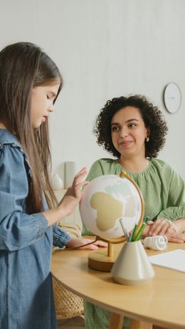 A child points at a globe on a desk while a person sits nearby smiling.