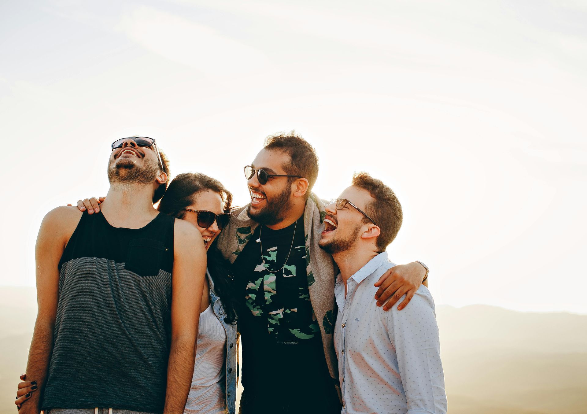 Four friends laughing and embracing outdoors in the sun.