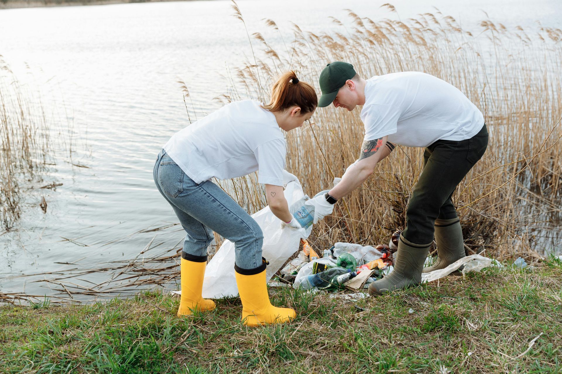 Two people picking up trash by a body of water; one wears yellow boots and the other a green cap.
