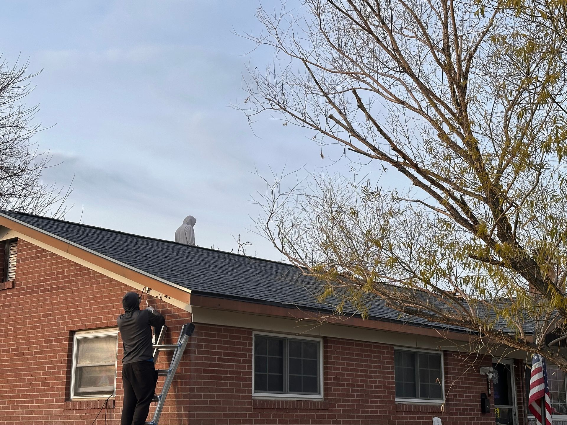 Person on ladder painting a brick house roofline beside leafless trees