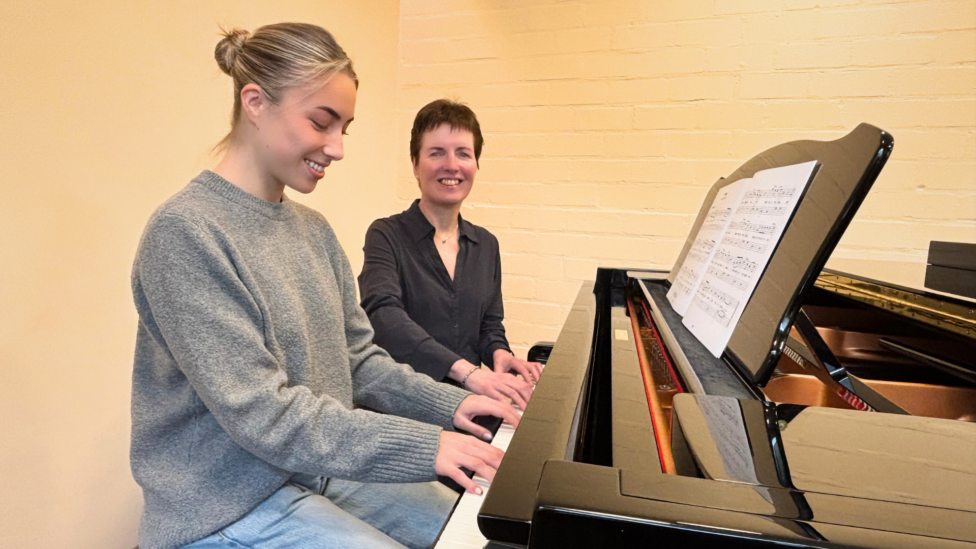 Een vrouw wijst naar bladmuziek voor een kind dat op een piano zit. Beiden glimlachen in een goed verlichte ruimte.