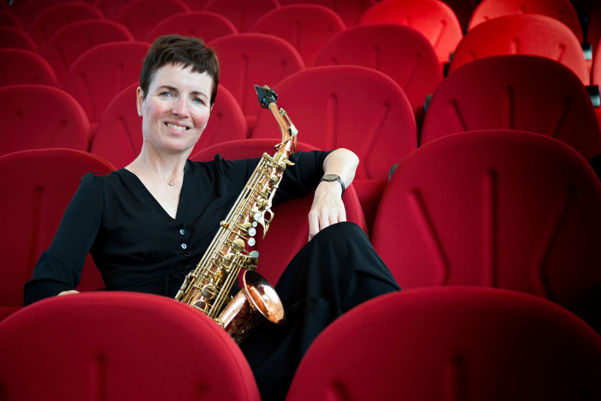 Vrouw met saxofoon, zittend op rode theaterstoelen.