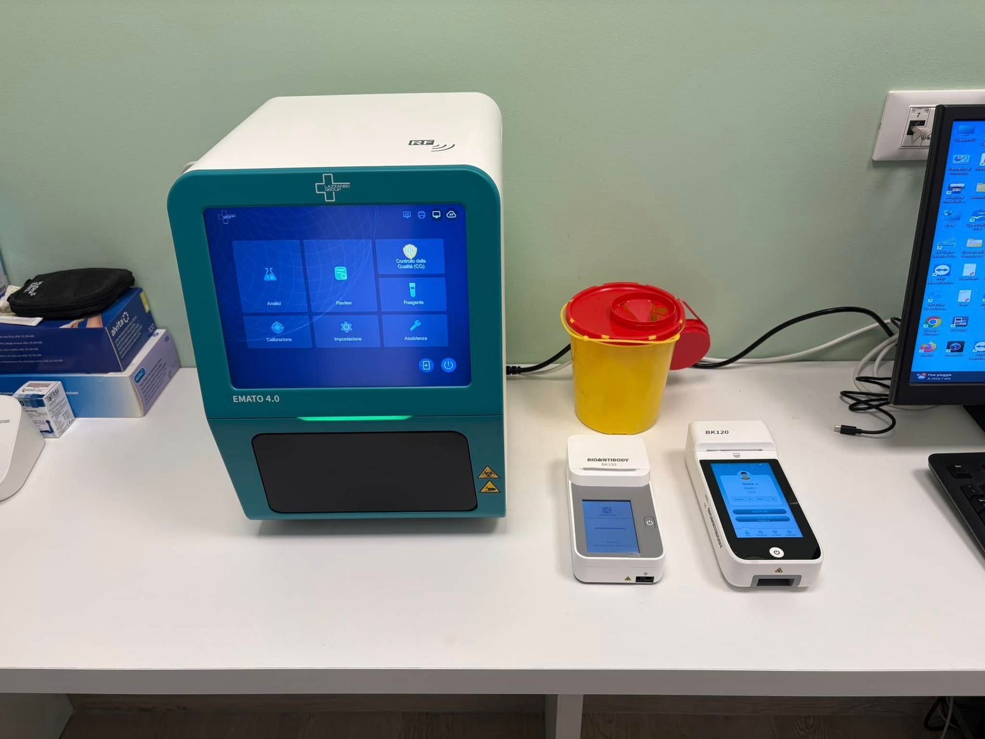 Banco da laboratorio con uno strumento color verde acqua, due piccoli dispositivi e una tazza giallo-rossa su un tavolo bianco.