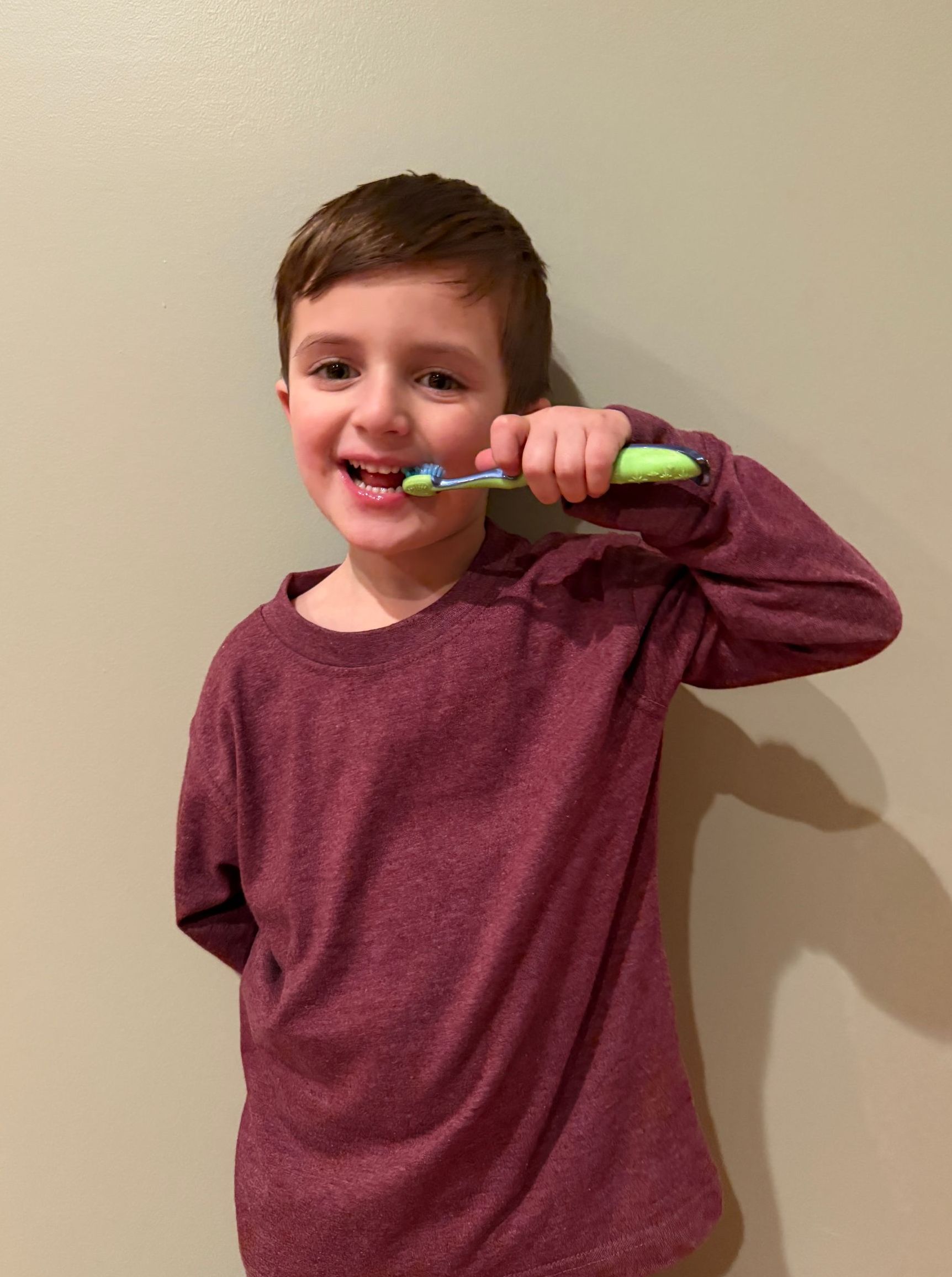 Boy brushing his teeth, smiling. He wears a maroon shirt and stands against a beige wall.