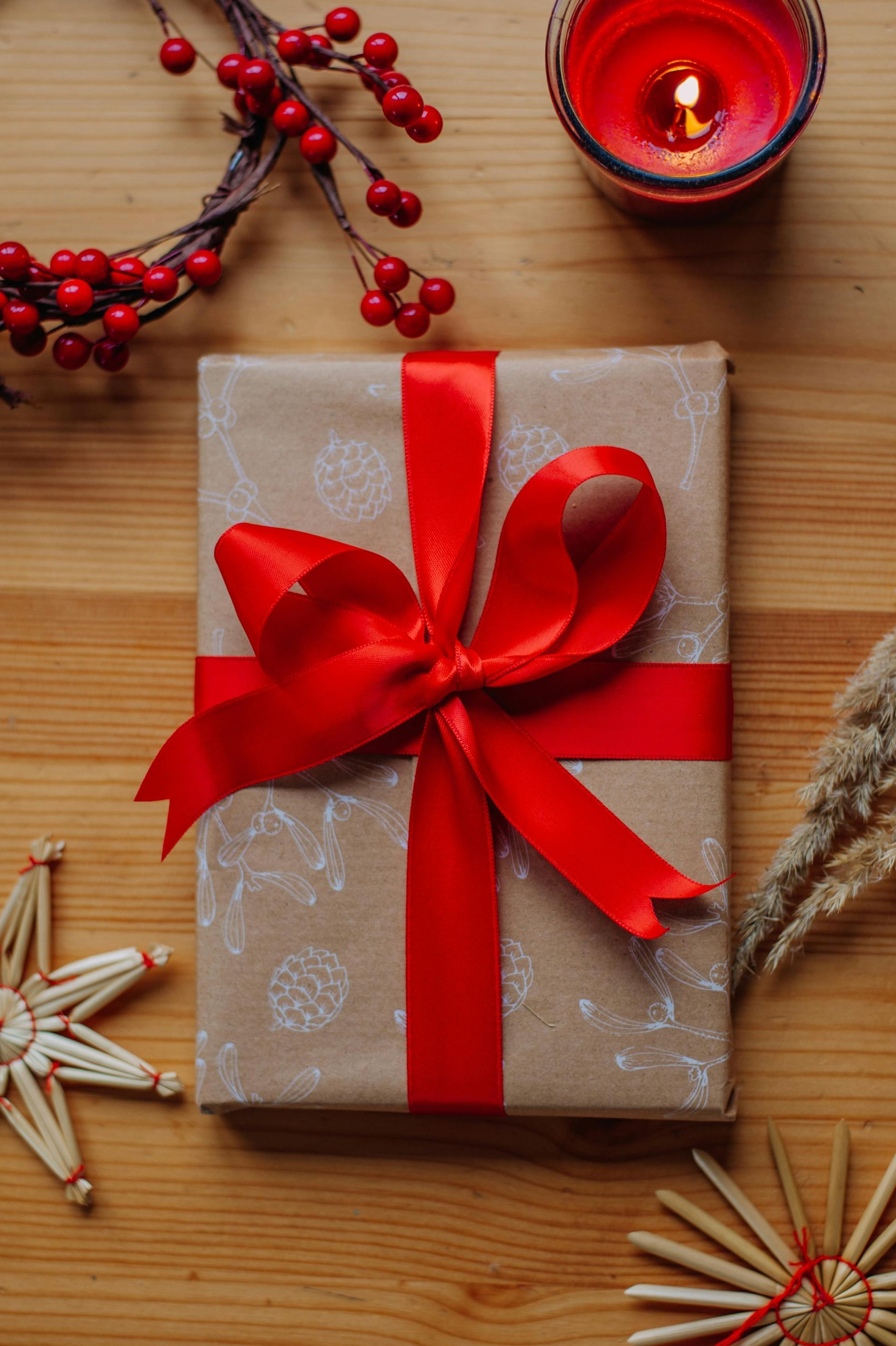 A gift wrapped in brown paper with a red ribbon on a wooden table, surrounded by holly berries, a candle, and straw stars.
