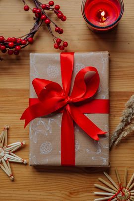 A gift wrapped in brown paper with a red ribbon on a wooden table, surrounded by holly berries, a candle, and straw stars.