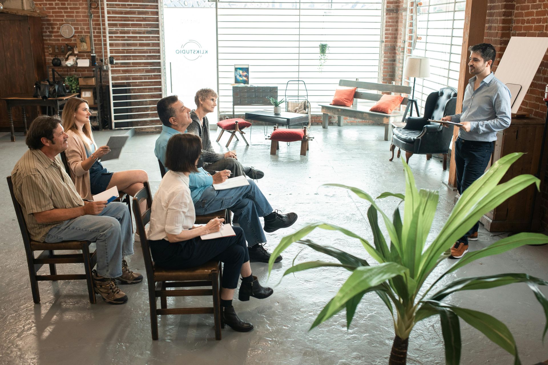 A group of people sit in chairs arranged in a circle, listening to a man standing and speaking in a sunlit loft workspace.