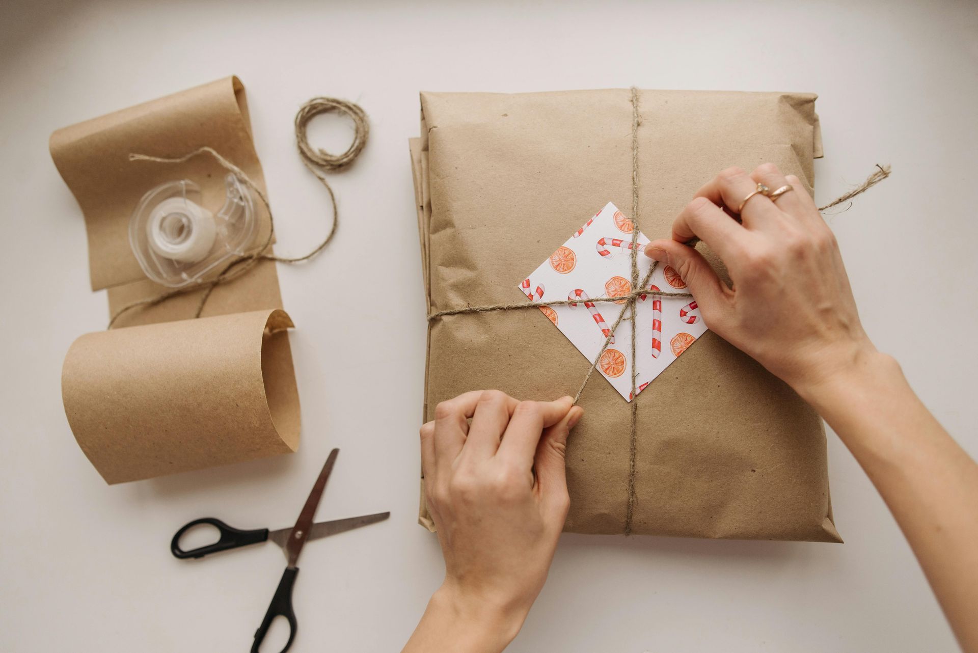 Hands wrapping a square gift in brown paper with twine and a patterned square tag, with scissors and tape nearby.
